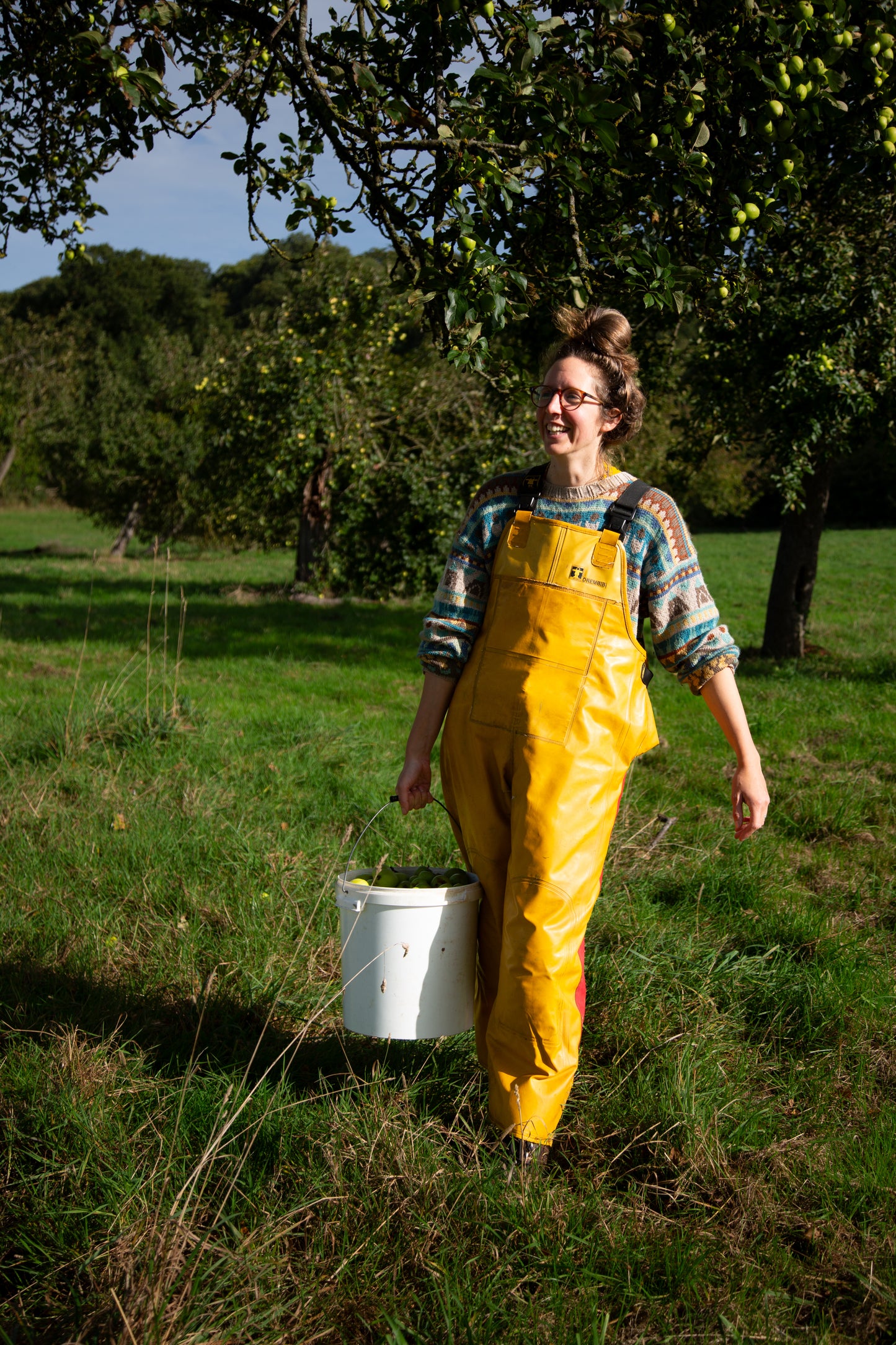 Person wearing yellow overalls holding a white bucket in a grassy field with trees in the background