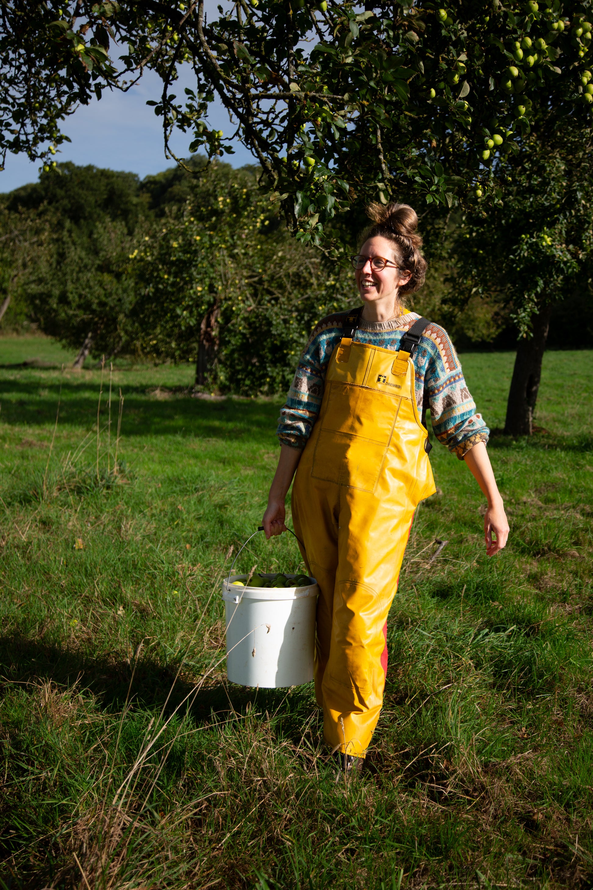 Person wearing yellow overalls holding a white bucket in a grassy field with trees in the background