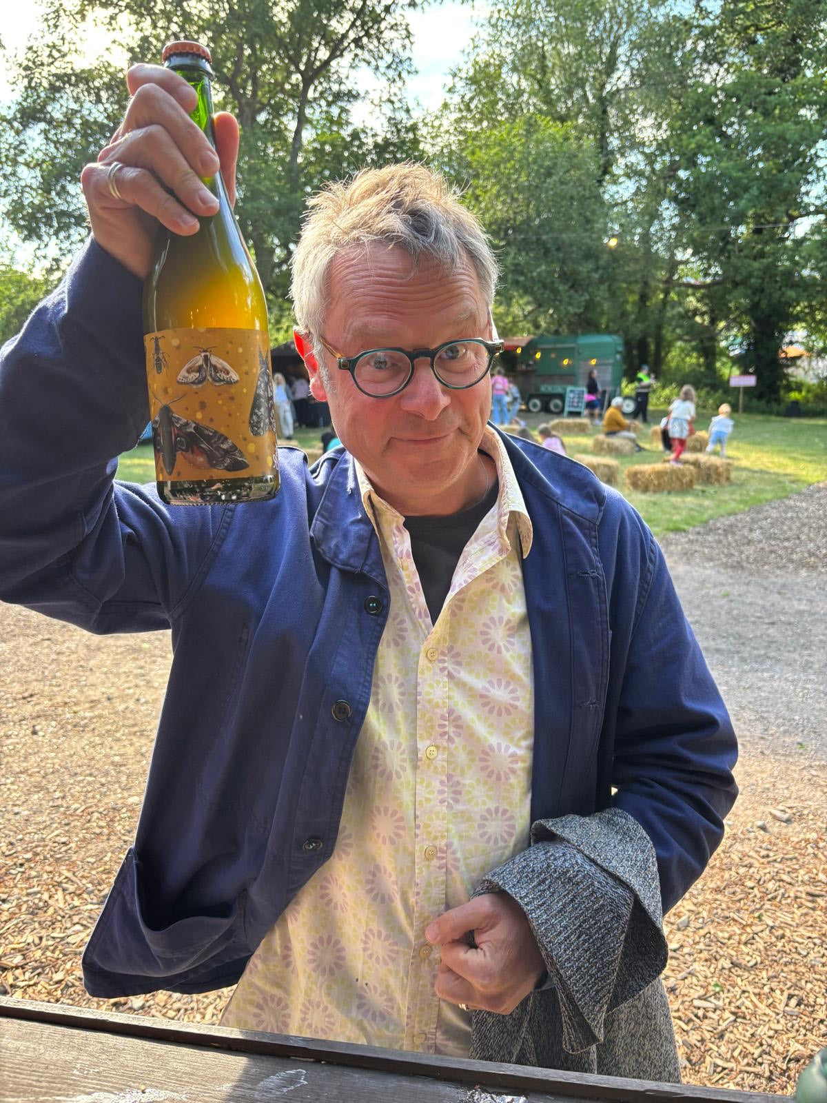 Hugh Fearnley-Whittingstall holding a bottle outdoors with trees and people in the background