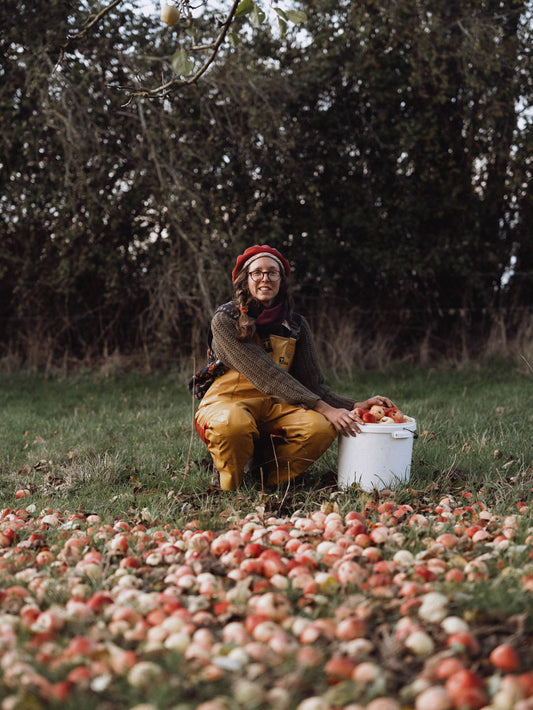 Person sitting on the ground surrounded by apples with a bucket nearby, in a natural setting.